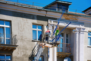 Two construction workers in bucket reconstructing facade of old building, worker install plastering mesh before plastering and painting works. Workers restore facade at height in lifting bucket.