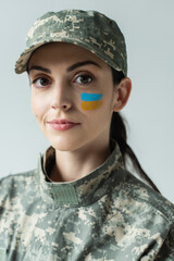 military woman with ukrainian flag painted on face looking at camera isolated on grey