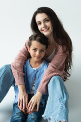 joyful brunette woman embracing daughter and looking at camera on grey background