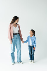 full length of woman and girl in jeans holding hands and smiling at each other on grey background