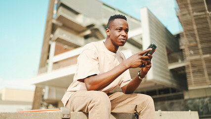 Young african student sits outside of university, uses cellphone, taps on smartphone screen