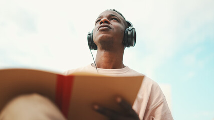 Young african student sitting outside of university and taking notes while study online with headphones