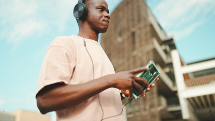 Young african student sitting outside of university wearing headphones, using phone, listening to music