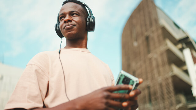 Young african student sitting outside of university wearing headphones, using phone, listening to music