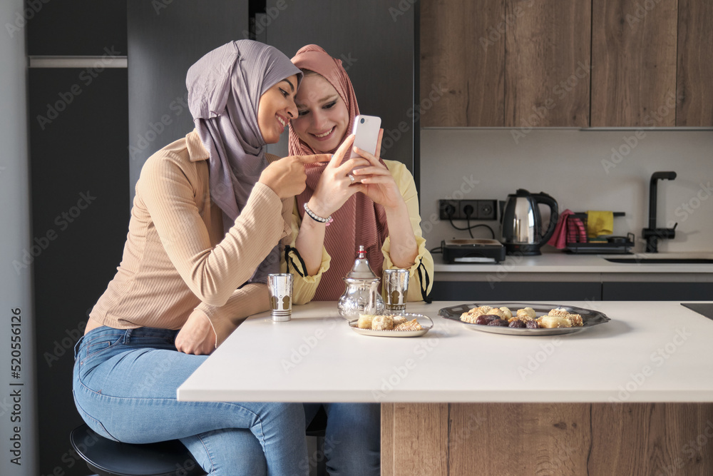 Wall mural two muslim women using the smartphone while having tea and arabic sweets at the kitchen table.