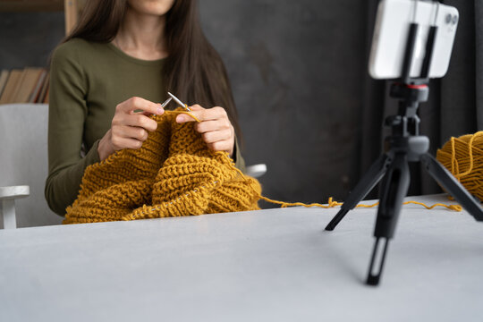 Young blogger shooting or videographing own needle crochet knitting by using a phone camera on a tripod. Woman blogger records a video lesson on knitting, close up of hands.
