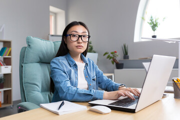 Distance Learning. A young Asian woman is a teacher, conducts online classes, lessons. He looks into the camera, explains, tells. Sitting in glasses and jeans at a table at a computer in a classroom