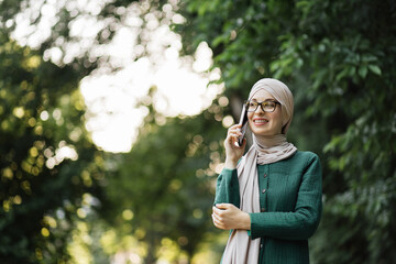 Portrait of smiling young muslim woman doing a phone call, taking on additional work during the break. Cheerful arabic woman in hijab using smart phone outdoor walking in park.