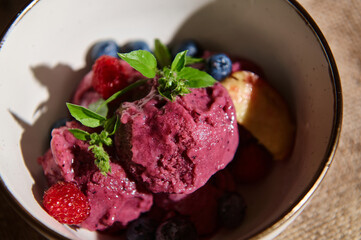 Raw vegan ice cream, sorbet with blueberries, raspberries, juicy peach slices and lemon basil leaves in a ceramic bowl.