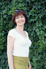 Woman with dark red hair stands and smiles in front of a wall covered in leaves. A woman lokking at the camera and smiling. Vertical portrait