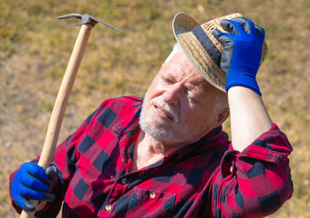 Porträt von einem älteren Mann mit Arbeitshandschuhen, Hut und Werkzeug. Er steht auf einem trockenen Acker im heißen Sonnenlicht und schwitzt.