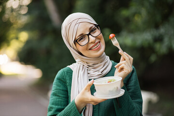 Happy positive muslim business woman eating healthy salad on a break standing over city park....