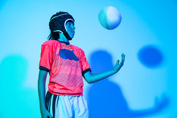 Portrait of junior male rugby player in sports uniform psoing with ball isolated on blue background in neon light. Sport, art, creativity concept