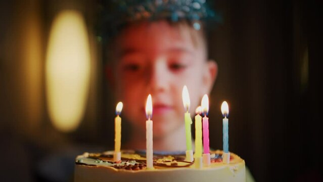 Close-up portrait in defocus of a little boy sitting at a table, mother to her son on his birthday lights candles on a cake on a birthday cake. Portrait child celebrating a birthday indoors at home.