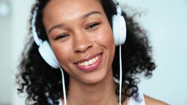 Cheerful curly haired woman listening music in headphones and looking at camera at home 
