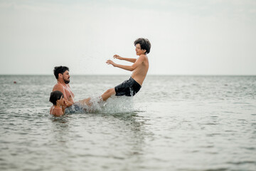 Dad swims with his son in the sea. The father throws his son in the water. The boy does a somersault in the sea. The family spends their summer vacation at the sea.