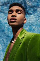 african american man with piercing posing in green blazer and beads near blue drape