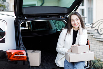 Girl sitting in the trunk of the car, holding boxes in his hands and smiling, looking at the camera. Concept of buying goods online and delivering them home
