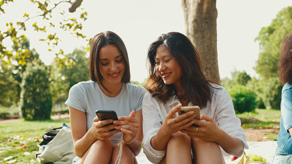 Young multi ethnic smiling women are watching videos on their phone. Girls laugh at picnic on...