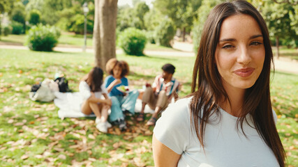 Naklejka premium Close-up of young smiling woman with long brown hair posing for the camera in the park . Picnic on summer day outdoors her friends sitting in distance blurred on background