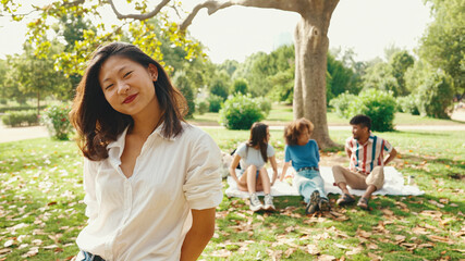Young Asian smiling woman with long brown hair wearing white t-shirt posing for the camera in the park . Picnic on summer day outdoors her friends sitting in distance blurred on background