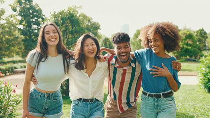 Happy multiethnic young people walk embracing on summer day outdoors. Group of friends are talking and laughing merrily while walking along path in city park