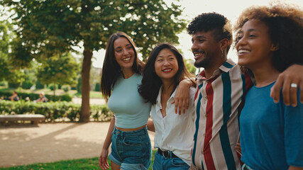 Happy multiethnic young people walk embracing on summer day outdoors. Group of friends are talking and laughing merrily while walking along path in city park