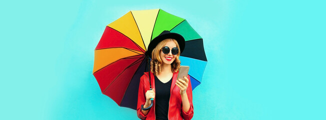 Autumn portrait of happy smiling young woman with smartphone holding colorful umbrella on blue...