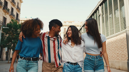 Happy multiethnic young people walk embracing on summer day outdoors. Group of friends are talking and laughing merrily while walking along on the street