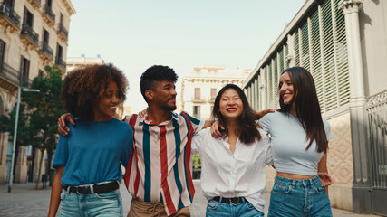Happy multiethnic young people walk embracing on summer day outdoors. Group of friends are talking and laughing merrily while walking along on the street