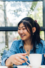 Positive Asian woman in casual clothes smiling while sitting at the table with a cup of hot drink.