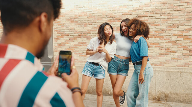 Happy, lovely multiethnic young people posing for the camera taking photo on summer day outdoors. Young man photographing smiling girls on cellphone camera