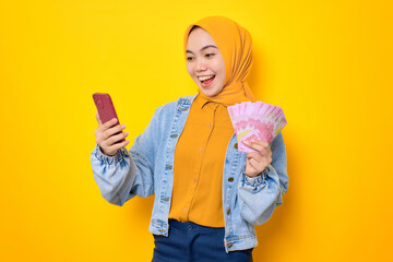 Fototapeta na wymiar Excited young Asian woman in jeans jacket holding mobile phone and rupiah banknotes, reacting to online win isolated over yellow background