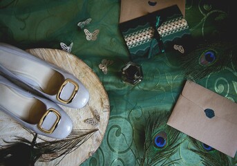 Top view of bridal shoes on a wooden surface next to peacock feathers and wedding invitations