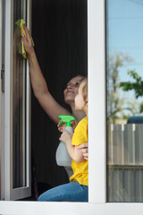 mother teaches her little daughter household chores, they wash the window in their house along with cleaning agent and yellow house cleaning rag