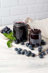 Homemade blueberry jam in glass jar at white table.
