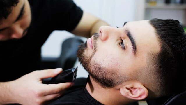 Barber electric trimmer shaves the beard of the client in the chair of the hairdressing salon.