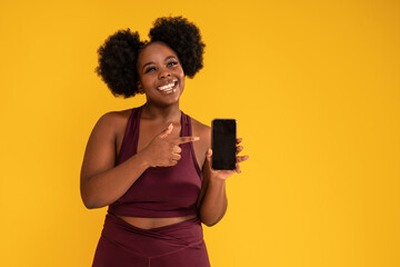 Smiling, happy, young girl with afro hairstyle posing on yellow studio background in sporty clothes, holding mobile phone in one hand showing new fitness app.