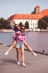 Happy young woman blond hung on back of her friend against the open background of city. Girls are smiling broadly  at camera.