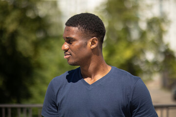 Portrait of young sportive smiling tall handsome African American black skin man looking away with a green tree on background, hot summer day in urban area. Horizontal plane. High quality photo.