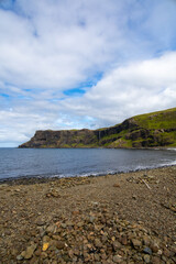 Fototapeta na wymiar Wide angle views of Talisker Bay, Isle of Skye, Scotland, with its rocky beach, black stones, green fields, and a waterfall at the end of beach. Scattered clouds on blue sky, summer scottish weather.