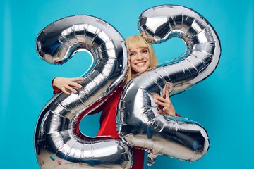 a fervently laughing, emotional woman in a red shirt stands on a blue background and holds inflatable balloons in the shape of the number twenty-two in silver color, hugging them with her hands