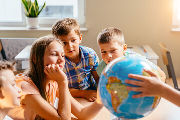Cheerful curious pupils playing with globe in classroom during geography lesson. Sun glare effect.