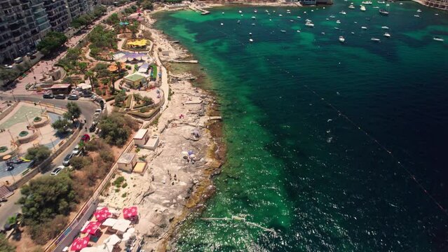 Drone flies over the blue water of Exiles Bay in Sliema. 4k aerial drone footage of beautiful beaches - Balluta Beach and Exiles Beach, Malta. People are sunbathing on the beach, playing in the sea.