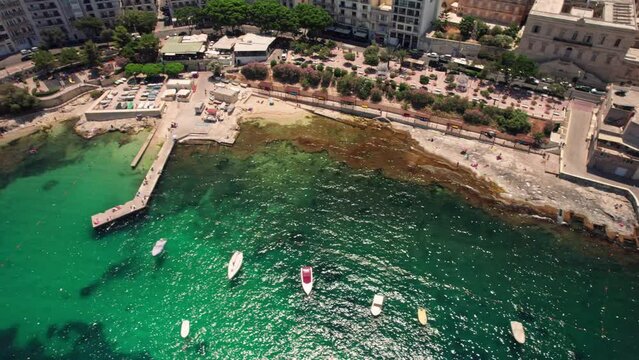 4k aerial drone footage of beautiful beaches - Balluta Beach and Exiles Beach, Malta. Drone flies over the blue water of Exiles Bay in Sliema. People are sunbathing on the beach, boats are in the sea.