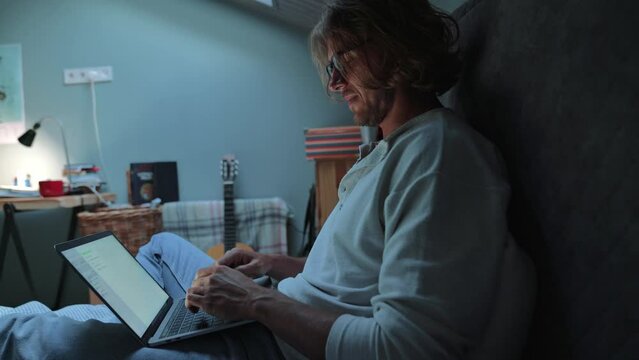 Cheerful blond man wearing eyeglasses working on laptop while sitting on the bed at home indoors