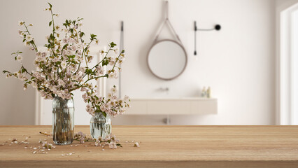 Wooden table, desk or shelf close up with branches of cherry blossoms in glass vase over blurred view of scandinavian white bathroom with washbasin, boho interior design concept