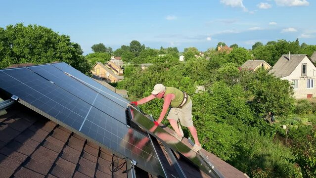 Worker carefully steps on roof to install new solar panels being secured with rope. DIY PV panels setup on bituminous tiles roof