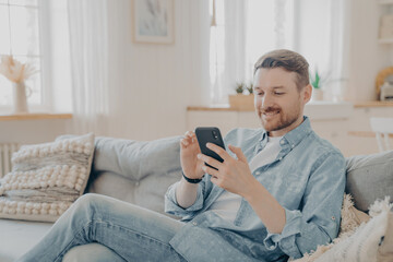 Happy smiling bearded man using smartphone device while relaxing on sofa