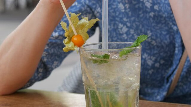 Summer cocktail. Mojito cocktail in a transparent glass glass with pieces of ice, a mint and lemon leaf, a woman drinks a cooling cocktail from a cocktail tube on the veranda of a summer cafe